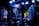 A band performs on stage with musicians playing electric guitars, surrounded by a crowd. The venue is dimly lit with blue and purple lighting creating a vibrant atmosphere, and spotlights highlight the performers and the audience.