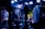A band performs on stage with musicians playing electric guitars, surrounded by a crowd. The venue is dimly lit with blue and purple lighting creating a vibrant atmosphere, and spotlights highlight the performers and the audience.