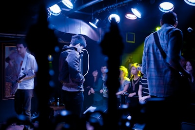 A band performs on stage with musicians playing electric guitars, surrounded by a crowd. The venue is dimly lit with blue and purple lighting creating a vibrant atmosphere, and spotlights highlight the performers and the audience.