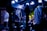 A band performs on stage with musicians playing electric guitars, surrounded by a crowd. The venue is dimly lit with blue and purple lighting creating a vibrant atmosphere, and spotlights highlight the performers and the audience.