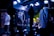 A band performs on stage with musicians playing electric guitars, surrounded by a crowd. The venue is dimly lit with blue and purple lighting creating a vibrant atmosphere, and spotlights highlight the performers and the audience.