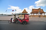A motorbike with a small attached passenger carriage rides along a road. To the right, there are two traditional-style pavilions with intricate roofs, and flags are visible. The scene is under a clear blue sky with scattered clouds, and it's set by a body of water.