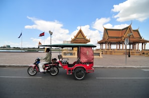 A motorbike with a small attached passenger carriage rides along a road. To the right, there are two traditional-style pavilions with intricate roofs, and flags are visible. The scene is under a clear blue sky with scattered clouds, and it's set by a body of water.