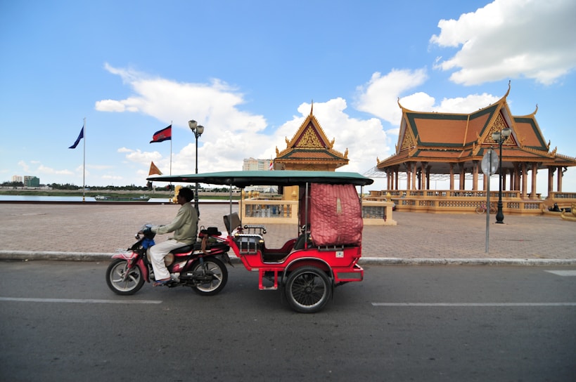 A motorbike with a small attached passenger carriage rides along a road. To the right, there are two traditional-style pavilions with intricate roofs, and flags are visible. The scene is under a clear blue sky with scattered clouds, and it's set by a body of water.