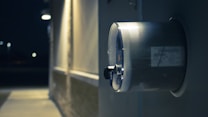 A close-up view of an outdoor electric meter mounted on a wall. The background is dimly lit, indicating it might be nighttime. The area surrounding the meter is illuminated by artificial light, creating a contrast between the bright and dark areas.
