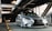 A sleek, silver Lexus car is parked in a modern parking structure with industrial metal beams overhead. The car has a sporty and polished appearance, with notable features including a black mesh grille and stylized headlights. The setting is dimly lit, with artificial lighting casting shadows on the surrounding architecture.