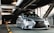 A sleek, silver Lexus car is parked in a modern parking structure with industrial metal beams overhead. The car has a sporty and polished appearance, with notable features including a black mesh grille and stylized headlights. The setting is dimly lit, with artificial lighting casting shadows on the surrounding architecture.