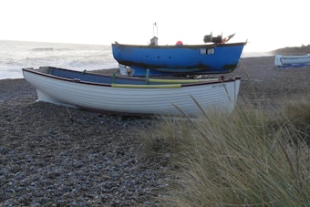 Two boats are resting on a pebble beach with some tall grass in the foreground. The nearest boat is white with a maroon trim and is named Bay Fisher, while the boat behind it is blue with a mix of green patches. The ocean is visible in the background with waves approaching the shore.