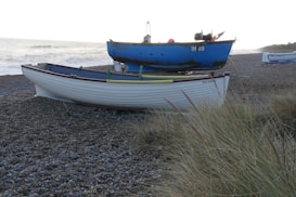 Two boats are resting on a pebble beach with some tall grass in the foreground. The nearest boat is white with a maroon trim and is named Bay Fisher, while the boat behind it is blue with a mix of green patches. The ocean is visible in the background with waves approaching the shore.