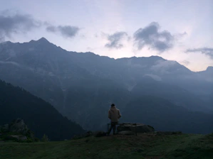 A weathered man in a worn coat stands alone on a misty hill, looking toward a distant, glowing city.