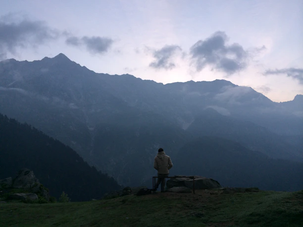 A weathered man in a worn coat stands alone on a misty hill, looking toward a distant, glowing city.