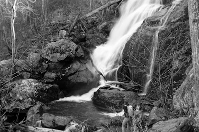 A waterfall cascades down rocky terrain into a small pool surrounded by natural vegetation. A person wearing a backpack stands near the water, capturing the scene with a camera. The monochrome palette emphasizes the textures of the rocks and water.