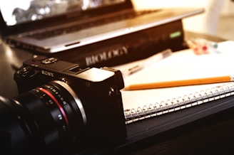 A close-up view of a camera placed on a table next to an open spiral notebook and a pencil. A laptop is partially visible in the background, with a textbook titled 'Biology' underneath it. Some colored pens are scattered on the table.