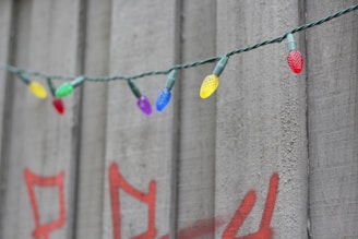 A string of colorful LED lights is hanging in front of a concrete wall. The wall features red graffiti, creating a contrast with the vibrant bulbs. The bulbs come in various colors including red, green, blue, yellow, and purple.
