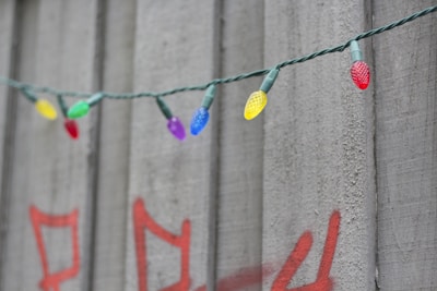 A string of colorful LED lights is hanging in front of a concrete wall. The wall features red graffiti, creating a contrast with the vibrant bulbs. The bulbs come in various colors including red, green, blue, yellow, and purple.