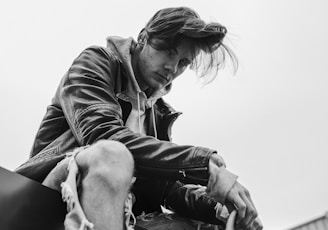 grayscale photo of man swearing leather jacket sitting on floor near building