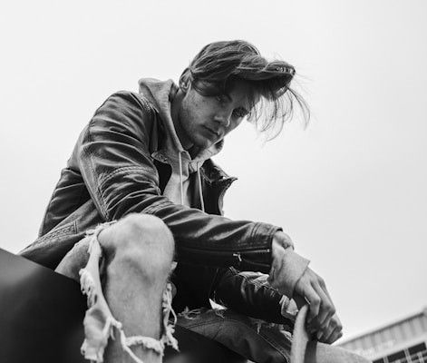 grayscale photo of man swearing leather jacket sitting on floor near building