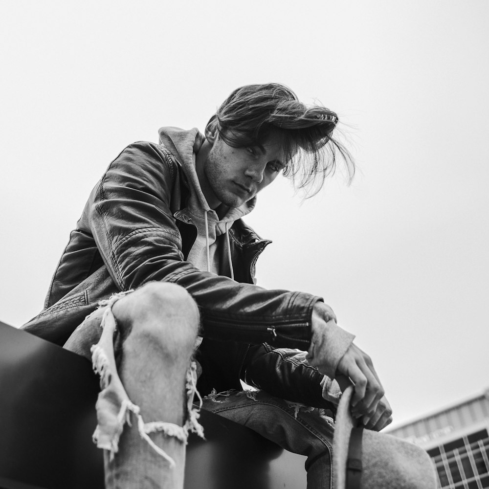 grayscale photo of man swearing leather jacket sitting on floor near building