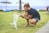 man squatting holding two foot of a white puppy on green sod at daytime