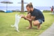 man squatting holding two foot of a white puppy on green sod at daytime