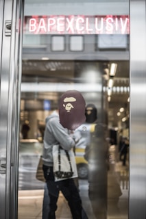 A fashion store entrance features a glass door with a reflective surface. A graphic logo of an ape head is prominently displayed on the glass. Behind the door, there are blurred figures of people walking and what appears to be shelves with products. Above the entrance, there is a neon sign with the word BAPECLUSIV, partially obscured by the door frame.