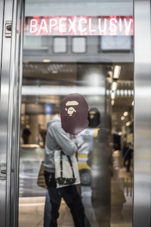 A fashion store entrance features a glass door with a reflective surface. A graphic logo of an ape head is prominently displayed on the glass. Behind the door, there are blurred figures of people walking and what appears to be shelves with products. Above the entrance, there is a neon sign with the word BAPECLUSIV, partially obscured by the door frame.