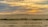 A group of people ride camels across a vast, sandy desert landscape under a sky painted with the warm hues of a setting sun. The horizon is lined with rolling dunes, and the open sky above is streaked with clouds reflecting the soft colors of sunset.