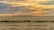 A group of people ride camels across a vast, sandy desert landscape under a sky painted with the warm hues of a setting sun. The horizon is lined with rolling dunes, and the open sky above is streaked with clouds reflecting the soft colors of sunset.