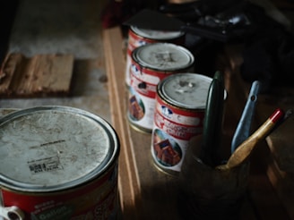 A collection of old and used paint cans is lined up on a wooden surface, accompanied by several paintbrushes in a container. The setup suggests a workspace for painting or DIY projects.