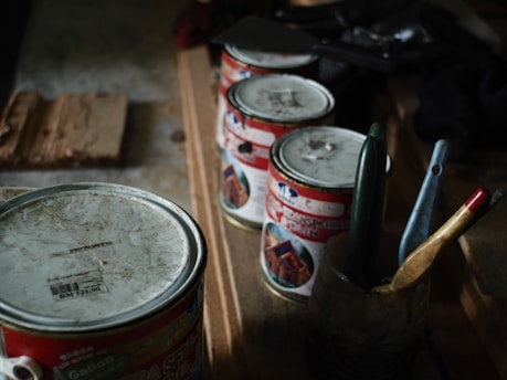 A collection of old and used paint cans is lined up on a wooden surface, accompanied by several paintbrushes in a container. The setup suggests a workspace for painting or DIY projects.