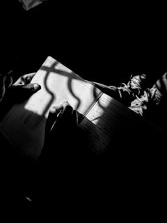 A person is writing or drawing in a notebook under the soft illumination of light, casting a shadow of a window frame onto the page. The image is black and white, emphasizing the contrast between light and shadow.