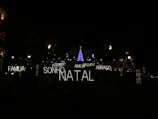A nighttime scene featuring festive illuminated words in Portuguese such as 'FAMILIA', 'SONHO', 'NATAL', 'AMOR', 'ABRACO', 'PAZ', and 'MAGIA'. In the background, a tall Christmas tree is adorned with blue and purple lights, surrounded by a dark environment with several streetlights and decorated trees visible.