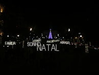 A nighttime scene featuring festive illuminated words in Portuguese such as 'FAMILIA', 'SONHO', 'NATAL', 'AMOR', 'ABRACO', 'PAZ', and 'MAGIA'. In the background, a tall Christmas tree is adorned with blue and purple lights, surrounded by a dark environment with several streetlights and decorated trees visible.