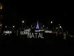 A nighttime scene featuring festive illuminated words in Portuguese such as 'FAMILIA', 'SONHO', 'NATAL', 'AMOR', 'ABRACO', 'PAZ', and 'MAGIA'. In the background, a tall Christmas tree is adorned with blue and purple lights, surrounded by a dark environment with several streetlights and decorated trees visible.
