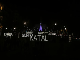 A nighttime scene featuring festive illuminated words in Portuguese such as 'FAMILIA', 'SONHO', 'NATAL', 'AMOR', 'ABRACO', 'PAZ', and 'MAGIA'. In the background, a tall Christmas tree is adorned with blue and purple lights, surrounded by a dark environment with several streetlights and decorated trees visible.