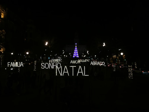 A nighttime scene featuring festive illuminated words in Portuguese such as 'FAMILIA', 'SONHO', 'NATAL', 'AMOR', 'ABRACO', 'PAZ', and 'MAGIA'. In the background, a tall Christmas tree is adorned with blue and purple lights, surrounded by a dark environment with several streetlights and decorated trees visible.