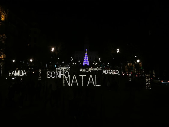 A nighttime scene featuring festive illuminated words in Portuguese such as 'FAMILIA', 'SONHO', 'NATAL', 'AMOR', 'ABRACO', 'PAZ', and 'MAGIA'. In the background, a tall Christmas tree is adorned with blue and purple lights, surrounded by a dark environment with several streetlights and decorated trees visible.