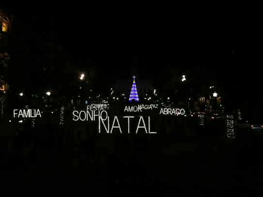 A nighttime scene featuring festive illuminated words in Portuguese such as 'FAMILIA', 'SONHO', 'NATAL', 'AMOR', 'ABRACO', 'PAZ', and 'MAGIA'. In the background, a tall Christmas tree is adorned with blue and purple lights, surrounded by a dark environment with several streetlights and decorated trees visible.