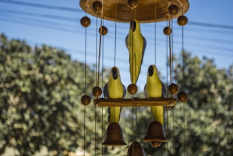 A wind chime with bird figures and bells hangs against a blurred background of trees and blue sky. The chime consists of three yellow bird shapes perched on a wooden frame, with spherical wooden beads and hanging bells creating a balanced arrangement.