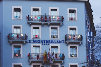 A light blue building with multiple windows and decorative black railings. The balconies are adorned with various colorful decorations and flags. The building has a sign reading 'Le Montbrillant' accompanied by four stars, indicating it is a hotel. A vertical sign on the side displays the word 'HOTEL'. The scene includes some trees partially visible in the foreground.