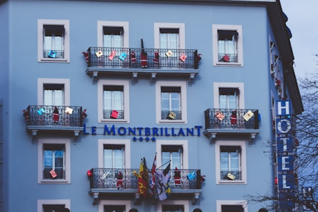A light blue building with multiple windows and decorative black railings. The balconies are adorned with various colorful decorations and flags. The building has a sign reading 'Le Montbrillant' accompanied by four stars, indicating it is a hotel. A vertical sign on the side displays the word 'HOTEL'. The scene includes some trees partially visible in the foreground.