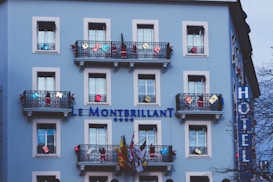A light blue building with multiple windows and decorative black railings. The balconies are adorned with various colorful decorations and flags. The building has a sign reading 'Le Montbrillant' accompanied by four stars, indicating it is a hotel. A vertical sign on the side displays the word 'HOTEL'. The scene includes some trees partially visible in the foreground.