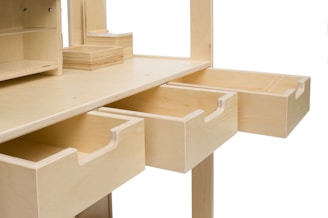 A wooden desk features multiple empty drawers that are extended outward. The desk is made from light-colored wood and has a minimalist design with clean edges and open shelves on top. The drawers have smooth pull handles carved into the wood.