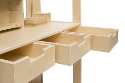 A wooden desk features multiple empty drawers that are extended outward. The desk is made from light-colored wood and has a minimalist design with clean edges and open shelves on top. The drawers have smooth pull handles carved into the wood.