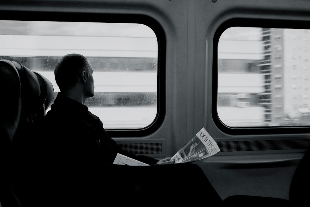 man inside train looking on window while holding newspaper grayscale photography