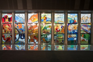 A series of colorful stained glass panels depicting various scenes. Each panel displays intricate designs featuring elements like trains, bridges, landscapes, workers, and people engaging in various activities. The scenes are rich in detail, with vibrant colors and reflective surfaces visible on the floor.