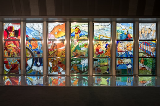 A series of colorful stained glass panels depicting various scenes. Each panel displays intricate designs featuring elements like trains, bridges, landscapes, workers, and people engaging in various activities. The scenes are rich in detail, with vibrant colors and reflective surfaces visible on the floor.