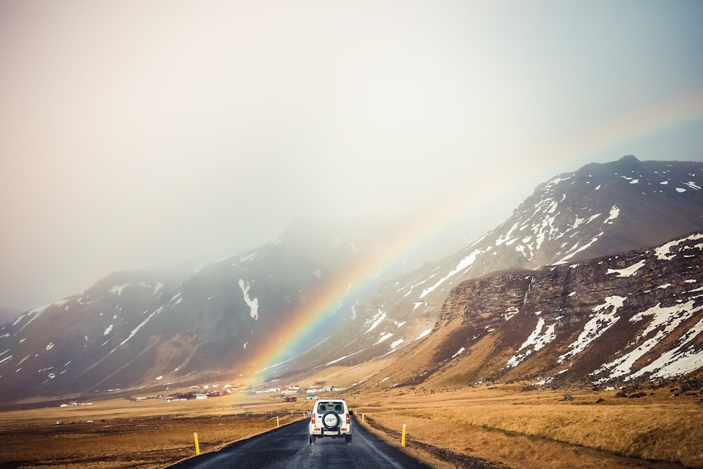 white car travelling near mountains with rainbow