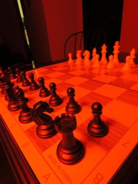 A chessboard is set up with black pieces on the left side and white pieces on the right, illuminated by a strong red light. The perspective is from one end of the board, giving prominence to the black pieces in the foreground. A knight and rook are clearly visible amongst the black pieces. The background is dimly lit with a shadowy chair in view.
