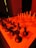 A chessboard is set up with black pieces on the left side and white pieces on the right, illuminated by a strong red light. The perspective is from one end of the board, giving prominence to the black pieces in the foreground. A knight and rook are clearly visible amongst the black pieces. The background is dimly lit with a shadowy chair in view.
